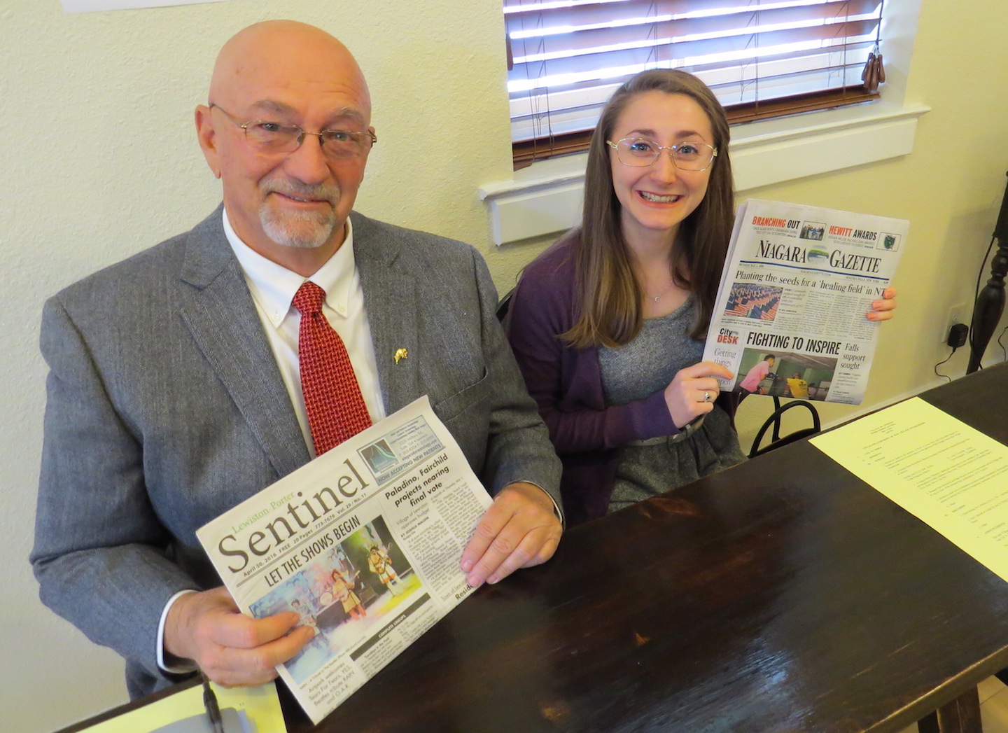 Bruce Sutherland, sitting in Joshua Maloni's seat, holds a copy of The Sentinel, while Joan McDonough, a reporter for the Niagara Gazette, hold a copy of her newspaper.