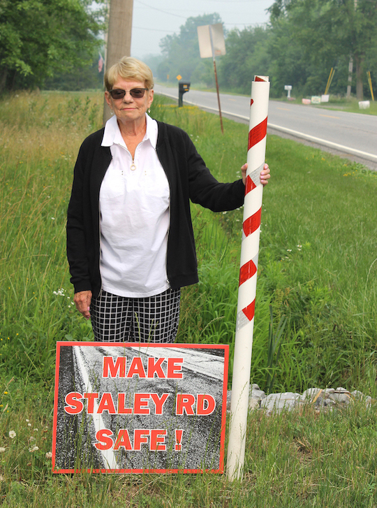 Alice Carlson stands by her driveway on Staley Road, where she has posted a sign calling for improvements to make the road safe. The signs dot yards on both sides along the length of Staley. She said cars and trucks move along the road so fast and frequently that they generate a forceful wind. She and other residents say that, with little to no shoulders on the roadside, it makes it hard to walk, bike or even retrieve the mail.